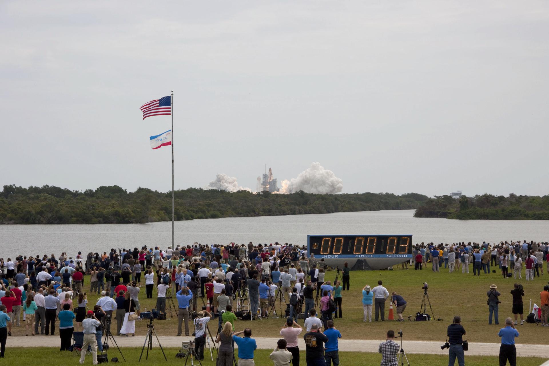 A crowd seen from behind watching a space shuttle lift off across the water, with a countdown clock beside them
